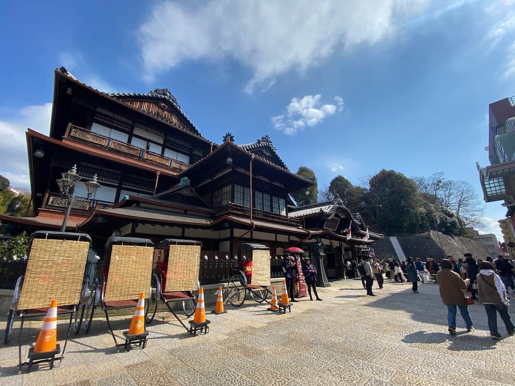 道後温泉本館の外観。趣のある木造建築の前には人力車が並び、観光客で賑わう様子が広がる。